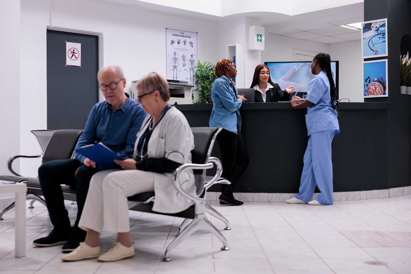 A waiting room with a man and a woman on chairs, reading a document. Two nurses and a receptionist converse at the front desk, creating a calm, professional atmosphere.