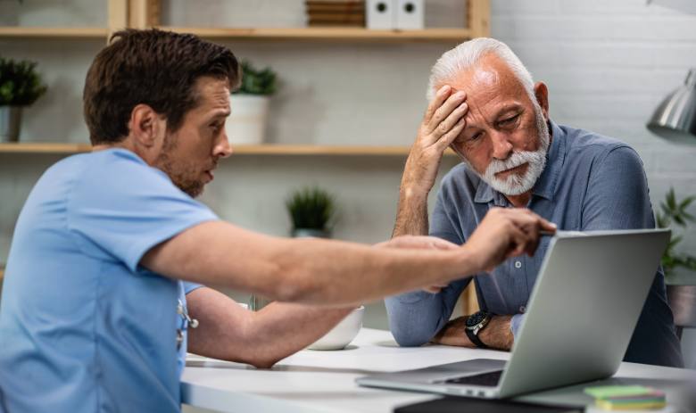 A doctor in scrubs discusses information on a laptop with an elderly man, who looks thoughtful and slightly concerned. They are in a well-lit office.