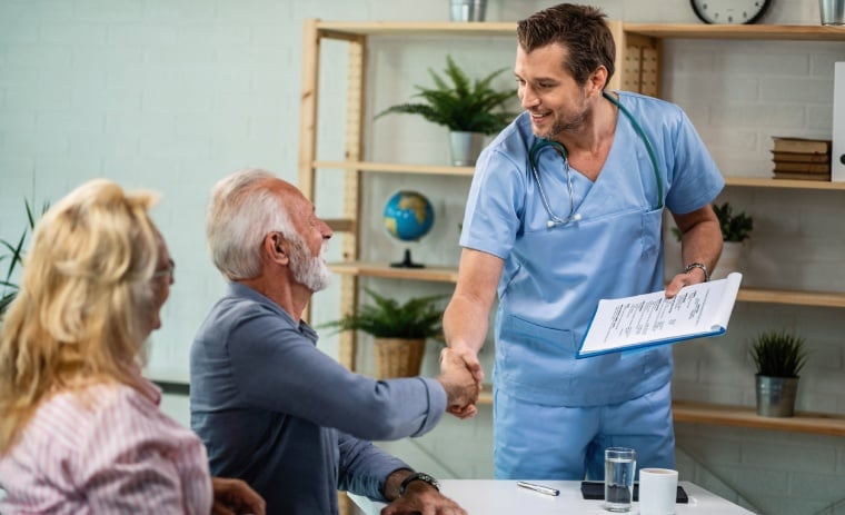 A male doctor in blue scrubs smiles warmly, shaking hands with an elderly man seated at a table. An elderly woman sits beside him. The mood is friendly.