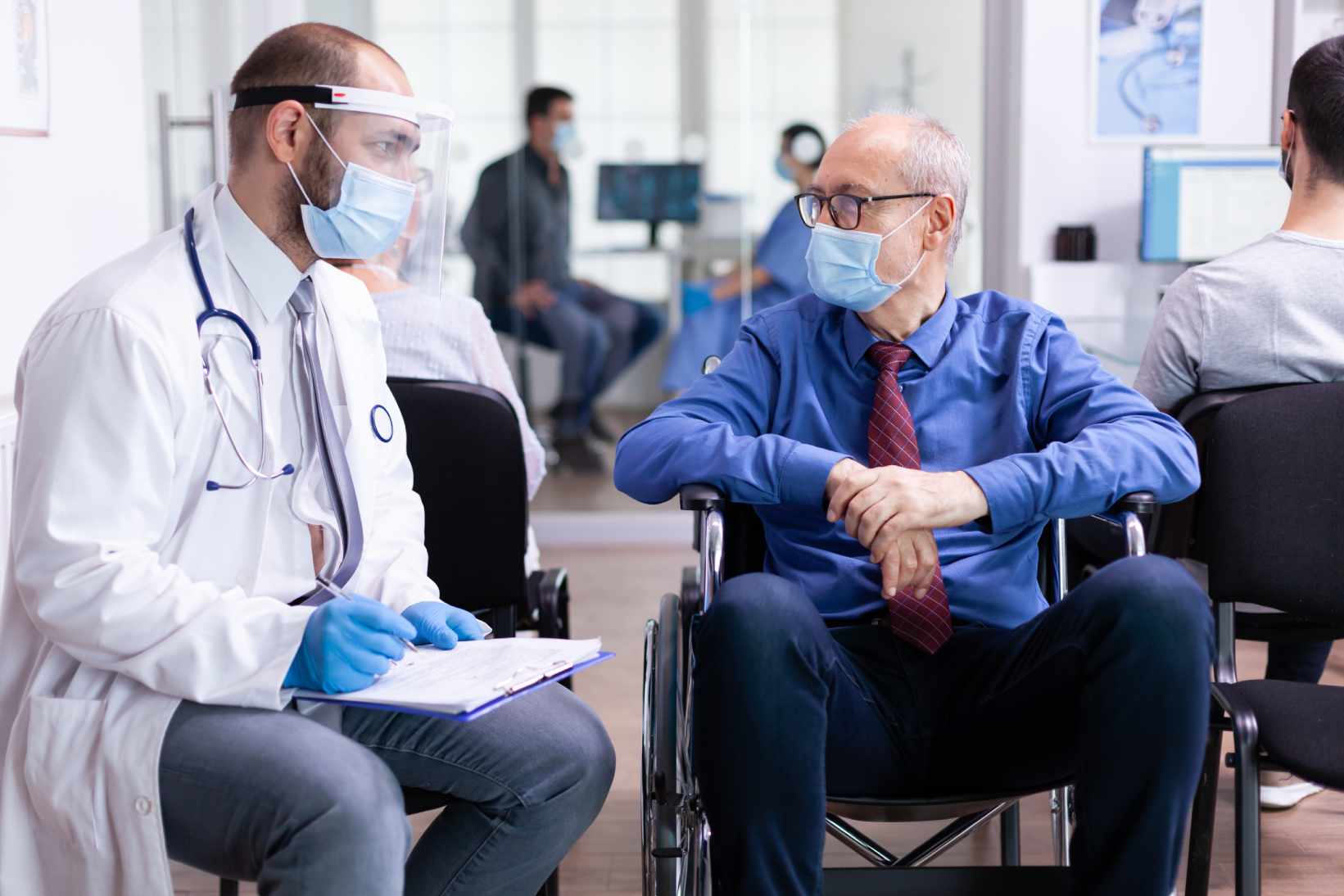 A doctor in a lab coat and face shield talks to an elderly patient in a wheelchair. Both wear masks. The setting is a bright medical office.