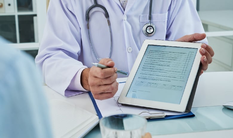A doctor in a white coat holds a tablet with a form displayed, discussing it with a patient. A stethoscope hangs around the doctor's neck, conveying professionalism.