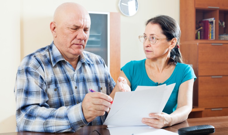 Elderly man in plaid shirt and woman in teal top sitting at a table, reviewing documents. They exhibit serious expressions, underlining focus and collaboration.